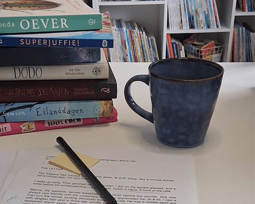 Werkplek van een kinderboekenschrijver met open manuscript vol correcties, laptop en stapel kinderboeken op een houten bureau bij het raam.