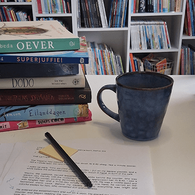 Werkplek van een kinderboekenschrijver met open manuscript vol correcties, laptop en stapel kinderboeken op een houten bureau bij het raam.