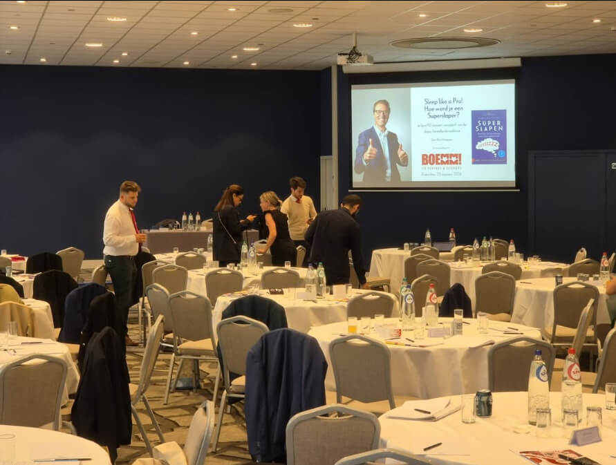 Conferentiezaal met deelnemers en een presentatie op het scherm over “Sleep like a Pro – Hoe word je een superslaper?”.